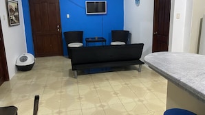 Una habitación con pared azul, sofá negro, dos sillas, una mesita, televisor y una puerta.