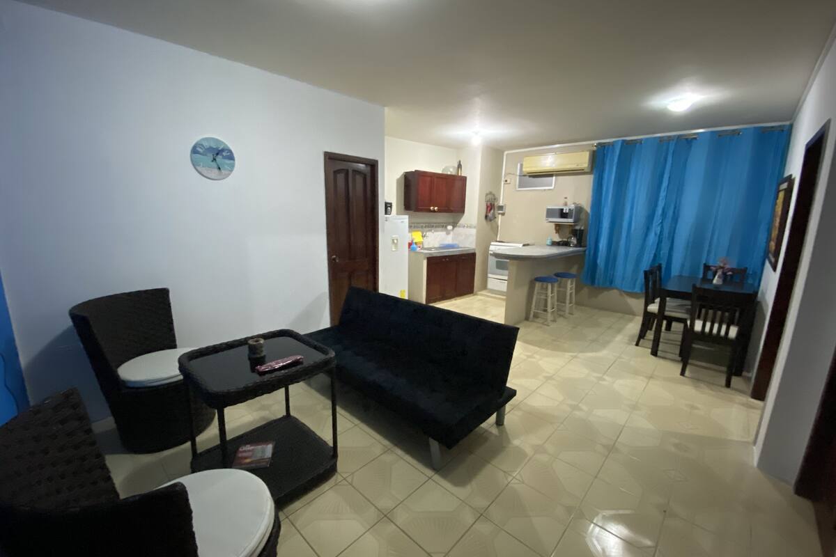 Un saló amb un sofà, una cadira i una taula de centre. Hi ha una zona de cuina amb nevera, fogó i tamborets. Es pot veure un rellotge a la paret.