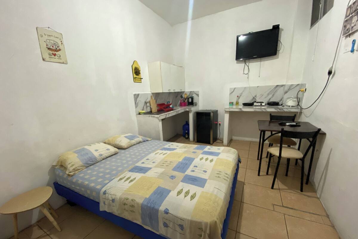 Habitació petita i senzilla amb un llit, escriptori, cadira i televisió penjada a la paret.