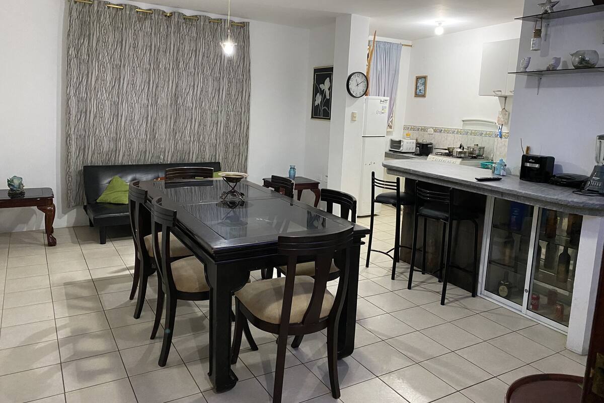 Zona de menjador amb taula rectangular i cadires, un sofà i una cuina que inclou nevera, microones i rentapelats.