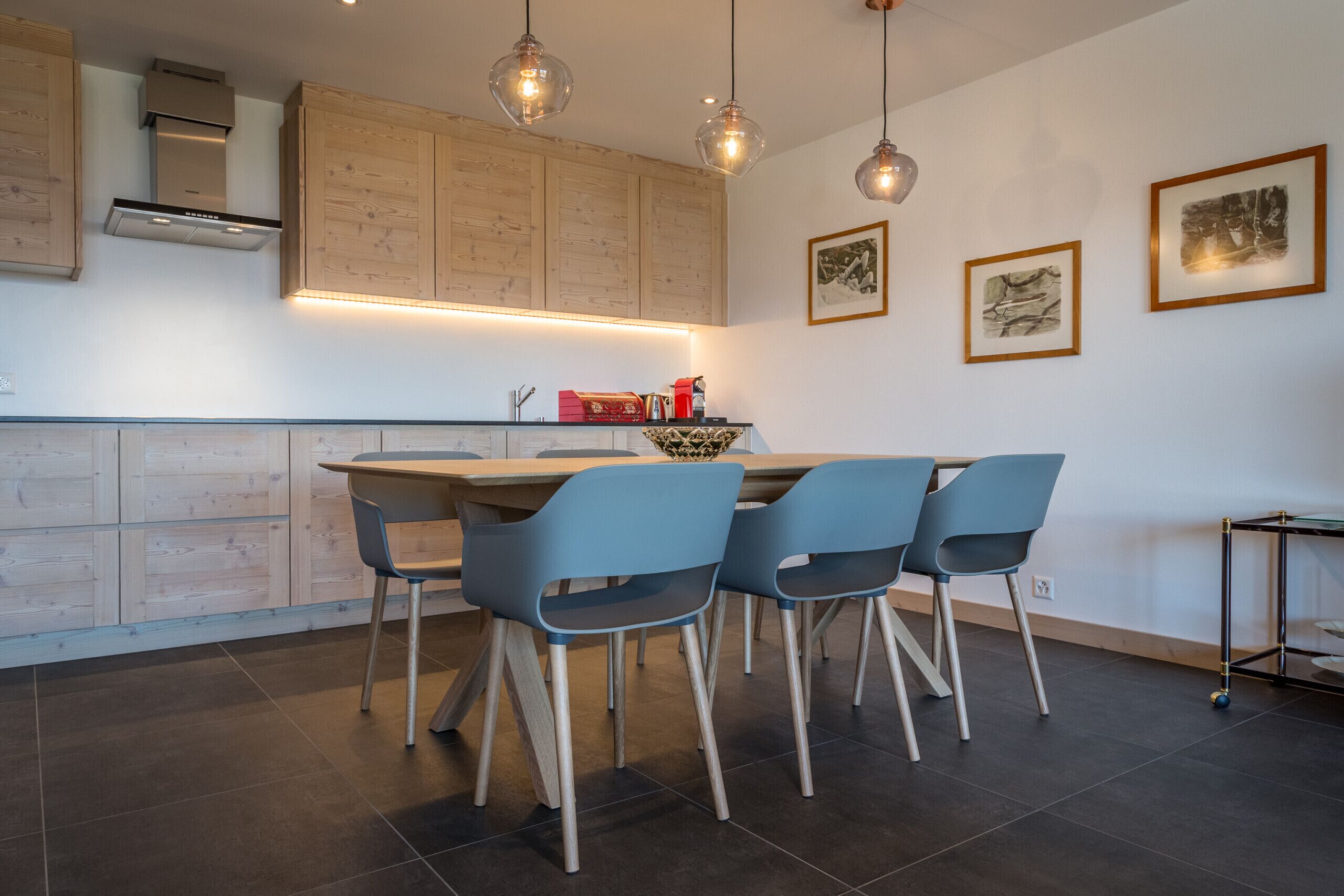 A modern kitchen with wooden cabinets, a central island with chairs, pendant lighting, and framed artwork on the wall.