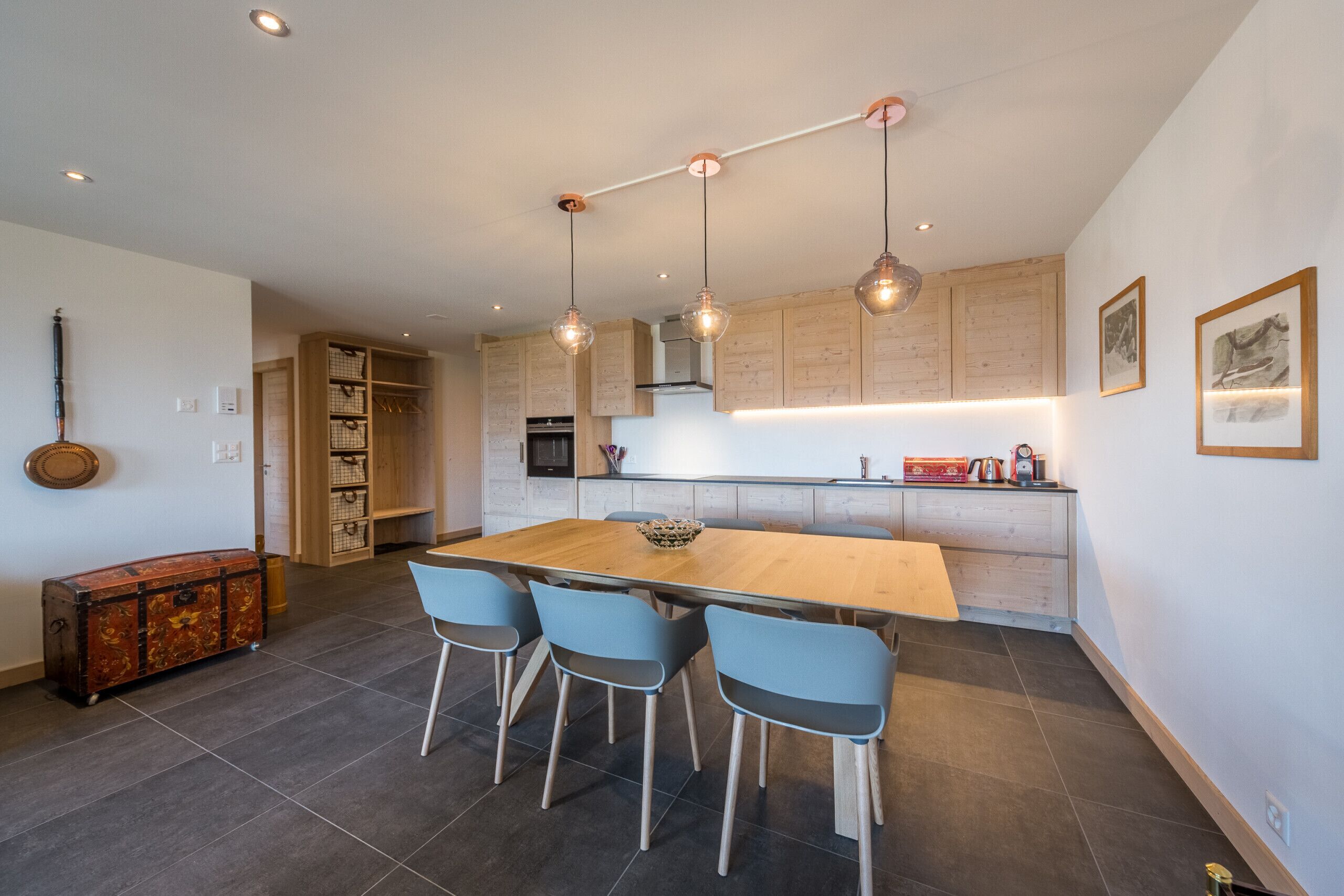 A modern kitchen with a wooden dining table, four chairs, pendant lights, and a built-in oven.
