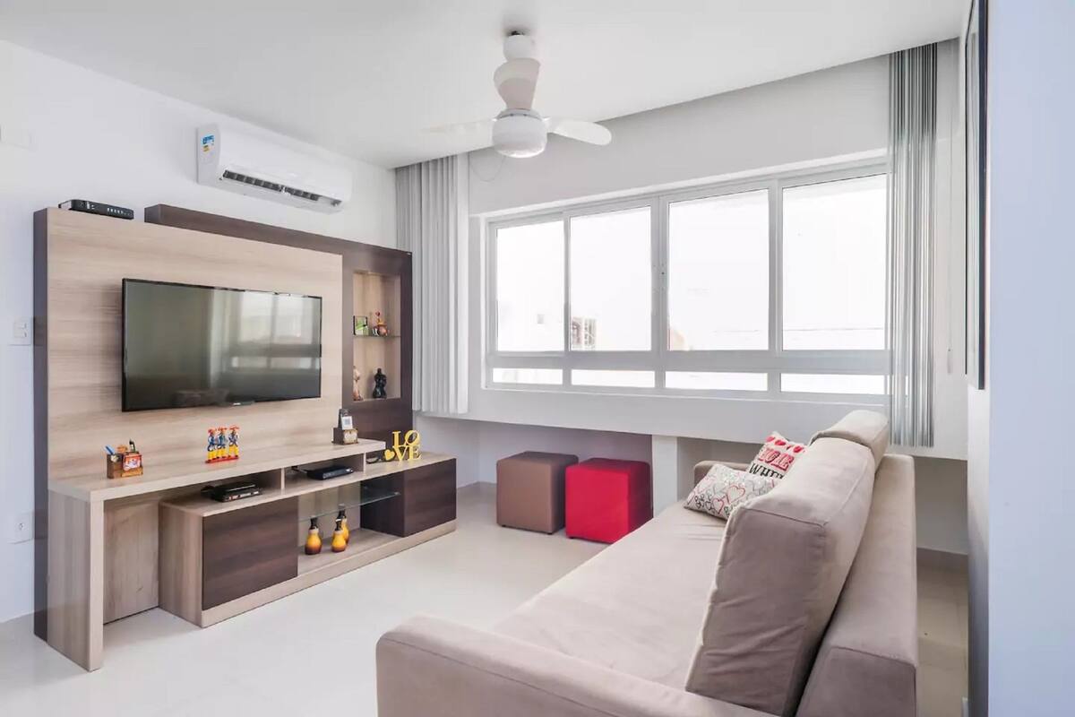 Una sala moderna con una televisión de pantalla plana, mueble de entretenimiento de madera, un reposapiés rojo, un sofá beige y un ventanal con cortinas.