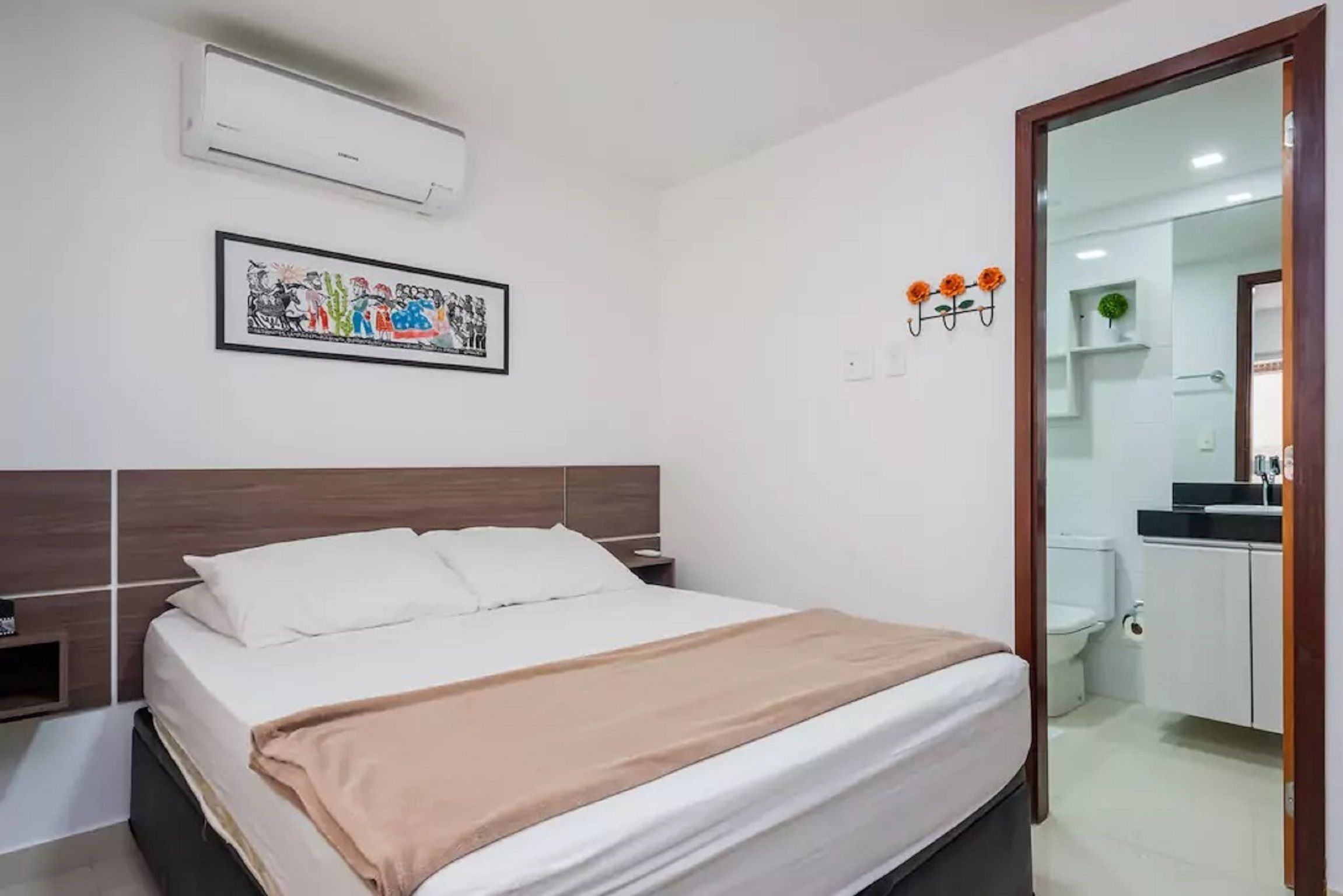 Habitación de hotel con cama, mesita de noche, unidad de aire acondicionado, un cuadro en la pared y un baño visible a través de una puerta abierta.