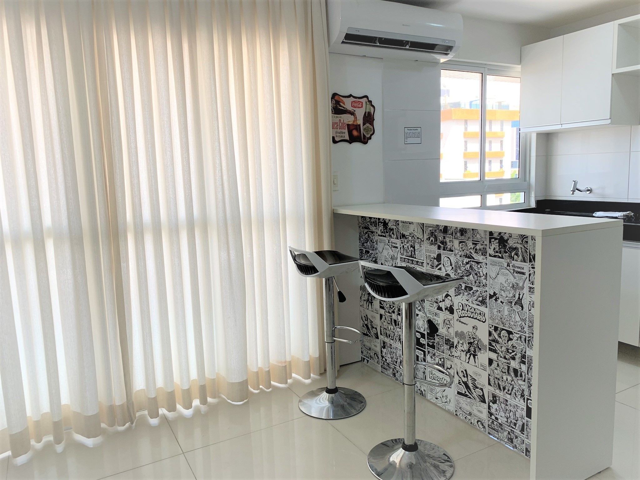 Una cocina moderna con armarios blancos, una barra con taburetes y un ventanal con cortinas.