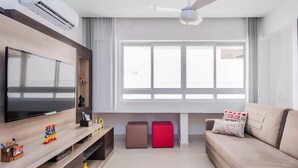 A modern living room with a flat-screen TV, a ceiling fan, a large window, a sofa, and two ottomans.