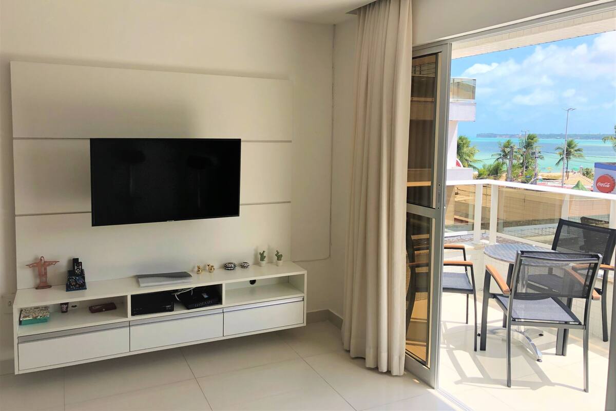 Una sala de estar moderna con un televisor de pantalla plana montado en la pared, un mueble blanco con varios objetos, y un balcón con puerta de vidrio que da a vistas del océano y palmeras.