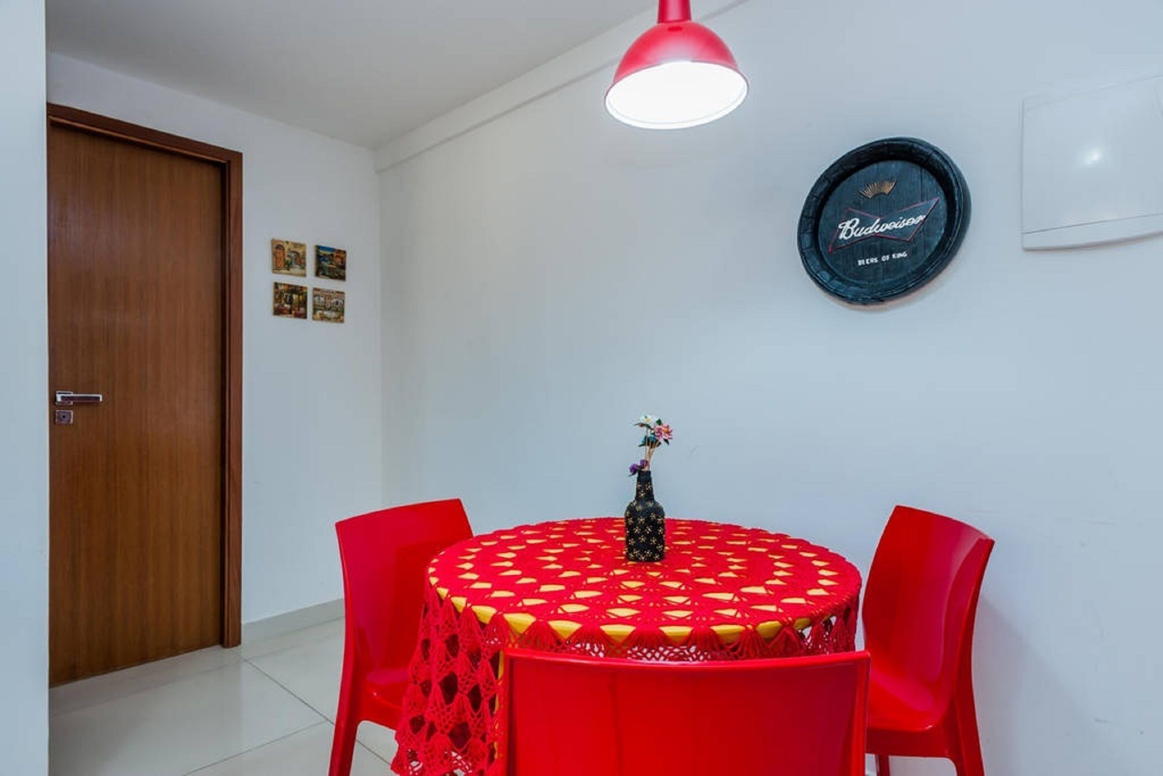 Zona pranzo con tavolo rotondo ricoperto da una tovaglia rossa, sedie rosse, logo Budweiser sulla parete e una porta in legno.