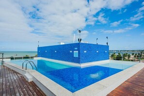 A rectangular swimming pool with a blue tiled deck, surrounded by a wooden deck and a metal railing. - Apartamento Tambau a Beira Mar (João Pessoa)