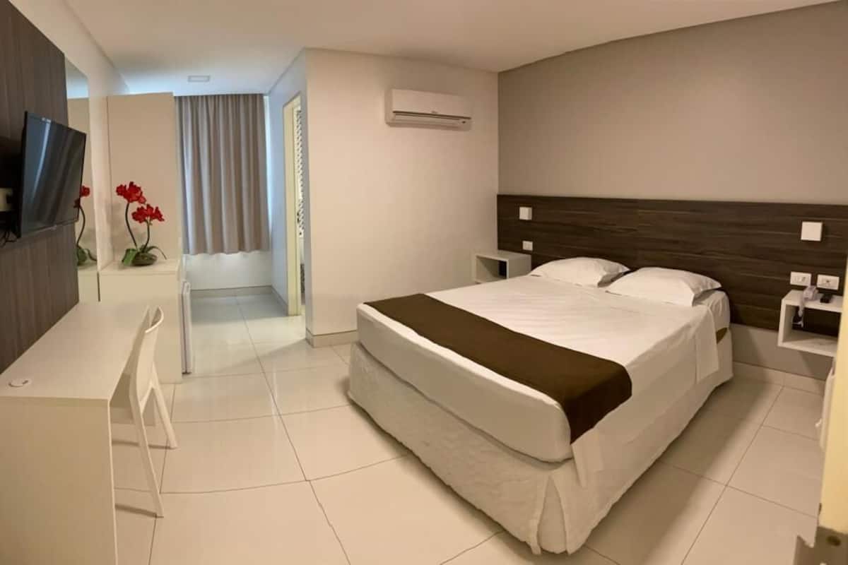 A hotel room with a bed, a desk, a chair, a TV, and an air conditioning unit.