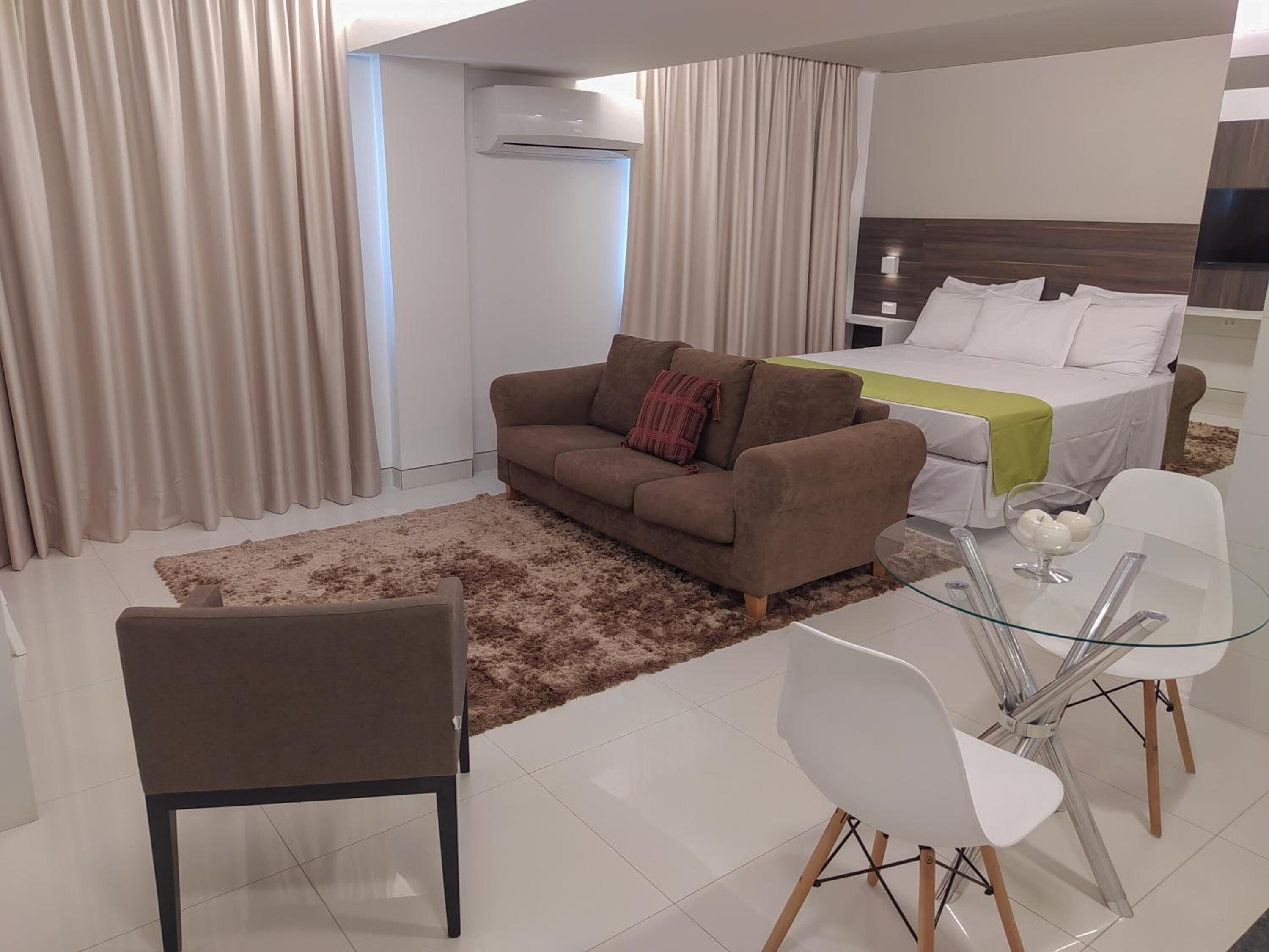 Una habitación de hotel moderna con un sofá marrón, una mesa de comedor de vidrio con sillas, una cama con ropa de cama blanca y un ventanal con cortinas.