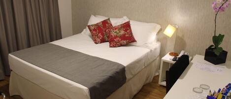 A neatly made bed with a grey bedspread, two red floral pillows, a bedside lamp, a small nightstand with a phone and a bowl of snacks, and a desk with a plant and stationery.
