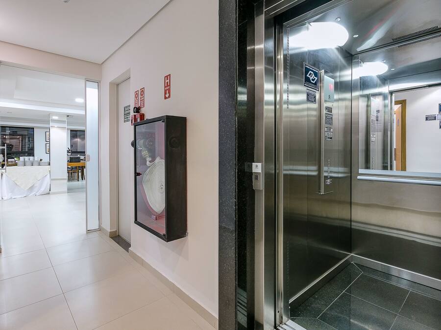 A modern elevator with stainless steel doors and a glass panel, a fire alarm on the wall, and a hallway leading to a dining area with a white tablecloth-covered table.