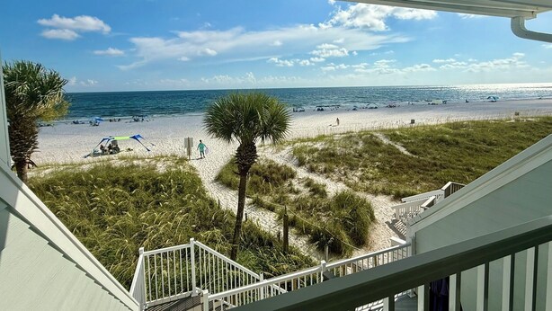 A beach view from a balcony with a railing, palm trees, and a sandy path leading to the beach.