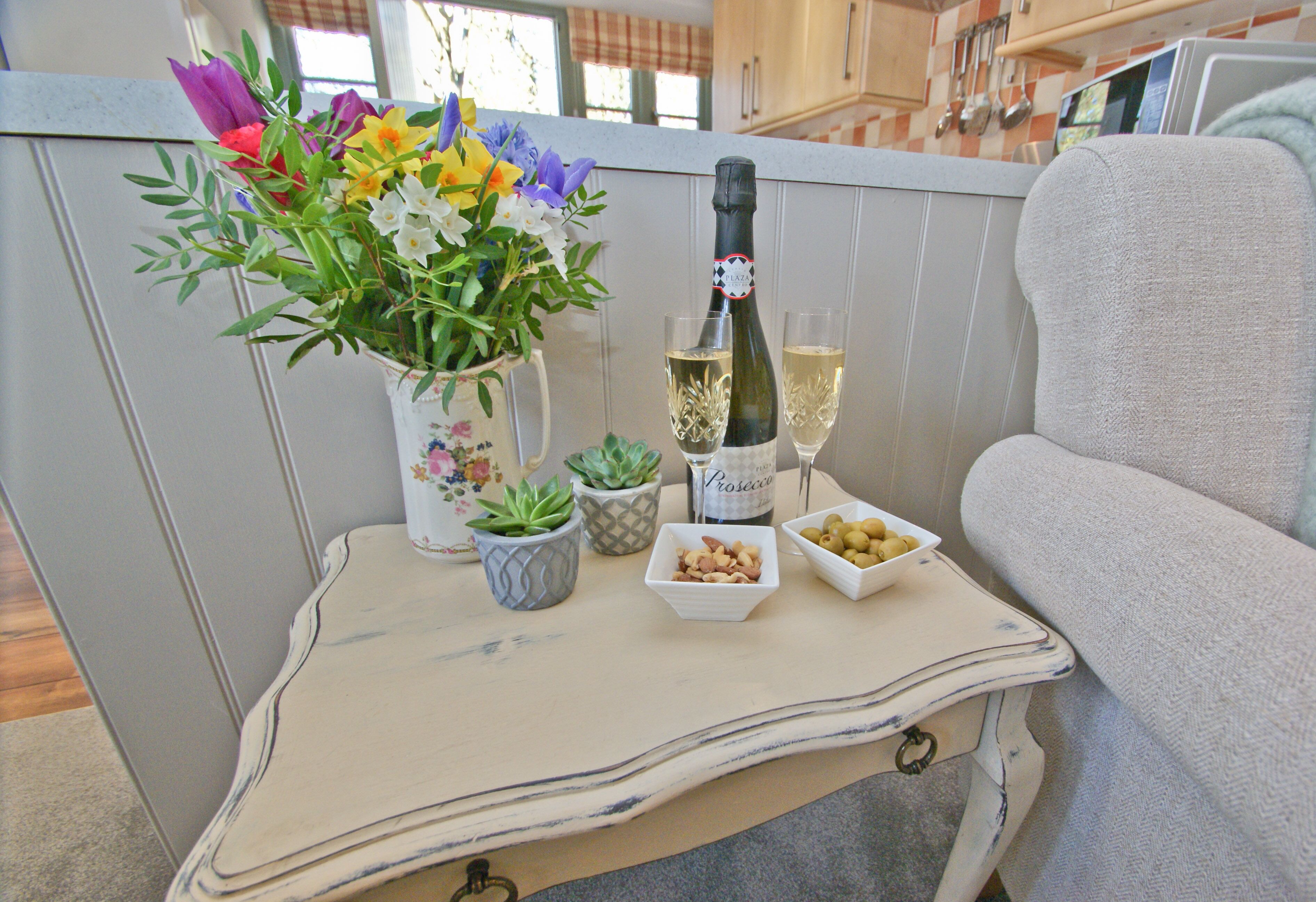 Una mesa con un jarrón de flores, una botella de champán, dos copas y recipientes pequeños con aperitivos.