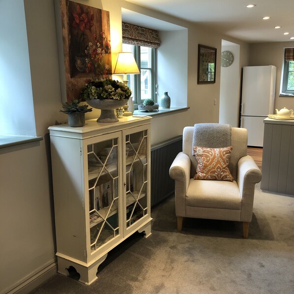 A cozy living room with a white armchair, a decorative cabinet, a painting, and a window with curtains.