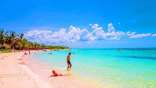 Una playa con agua turquesa cristalina, gente nadando y palmeras.