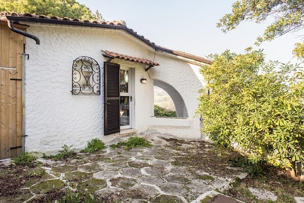 A rustic building with a white exterior, a wooden door, a window with decorative ironwork, and a stone-paved entrance.