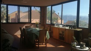 Un coin repas avec une table dressée pour deux personnes, de grandes fenêtres offrant une vue sur les bâtiments et les montagnes, et une petite table d’appoint agrémentée d’objets décoratifs.