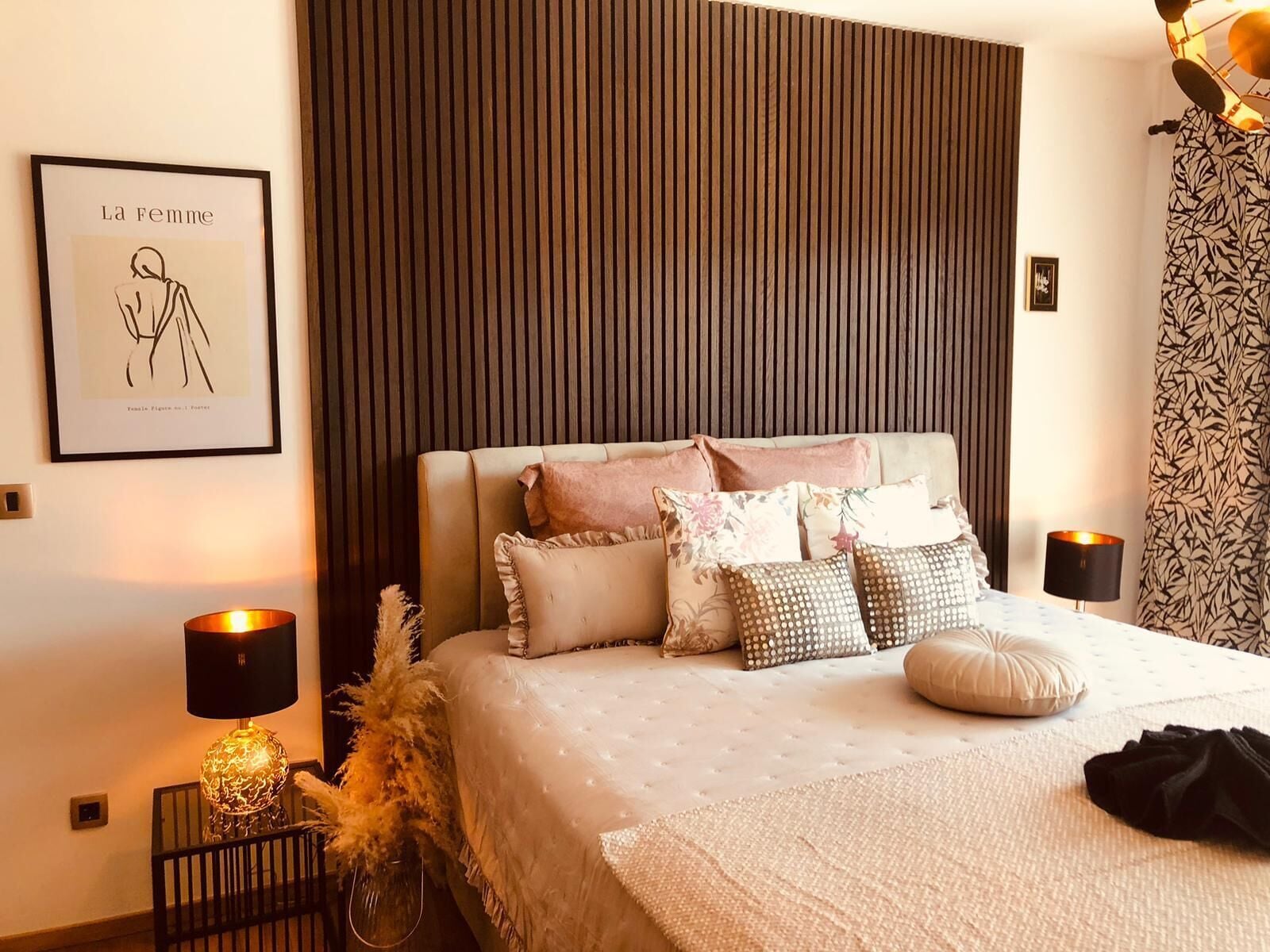 A neatly arranged bedroom with a large bed, multiple pillows, a framed artwork on the wall, and a decorative lamp.