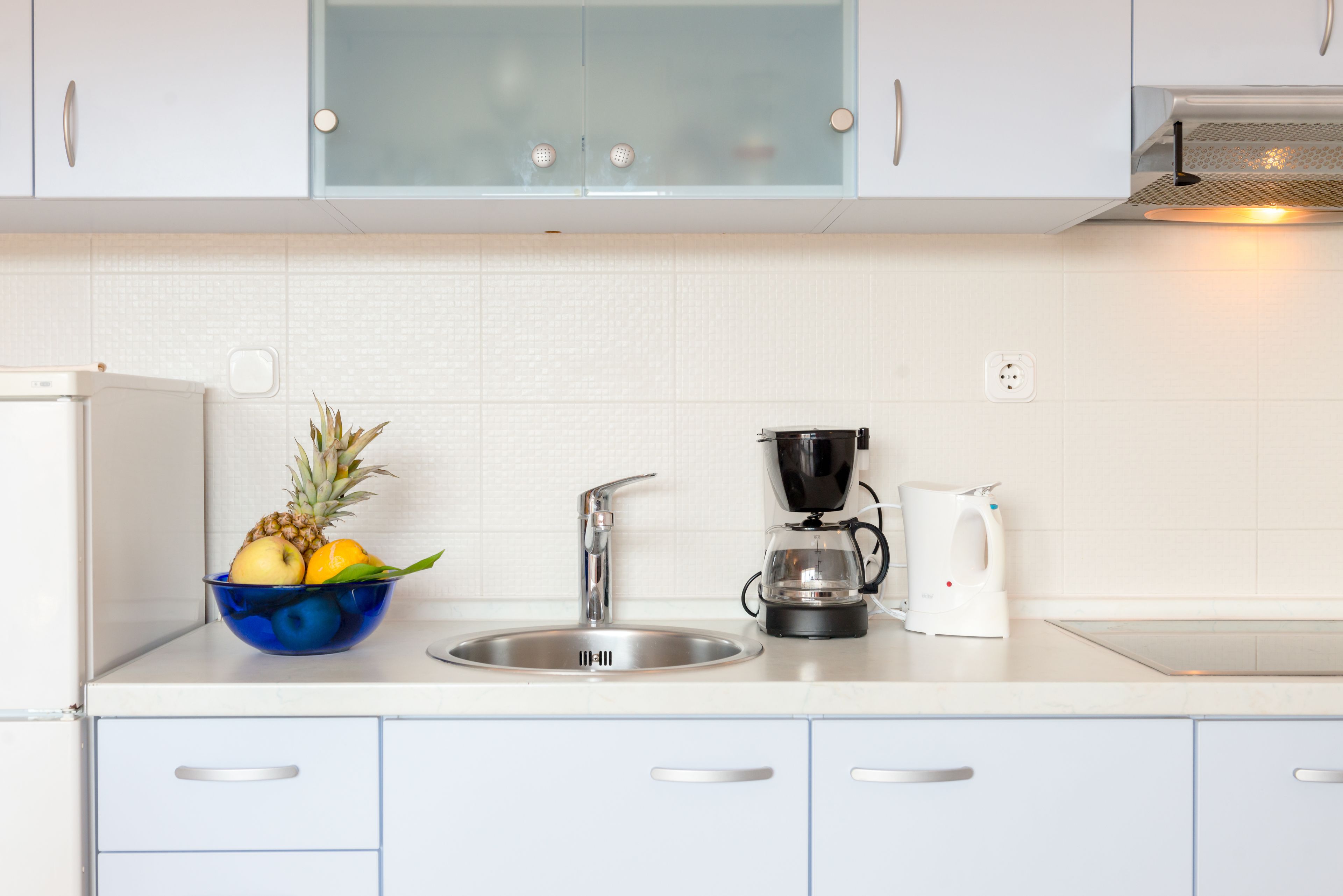 A modern kitchen with a white countertop, a stainless steel sink, a coffee maker, and a bowl of fruit.