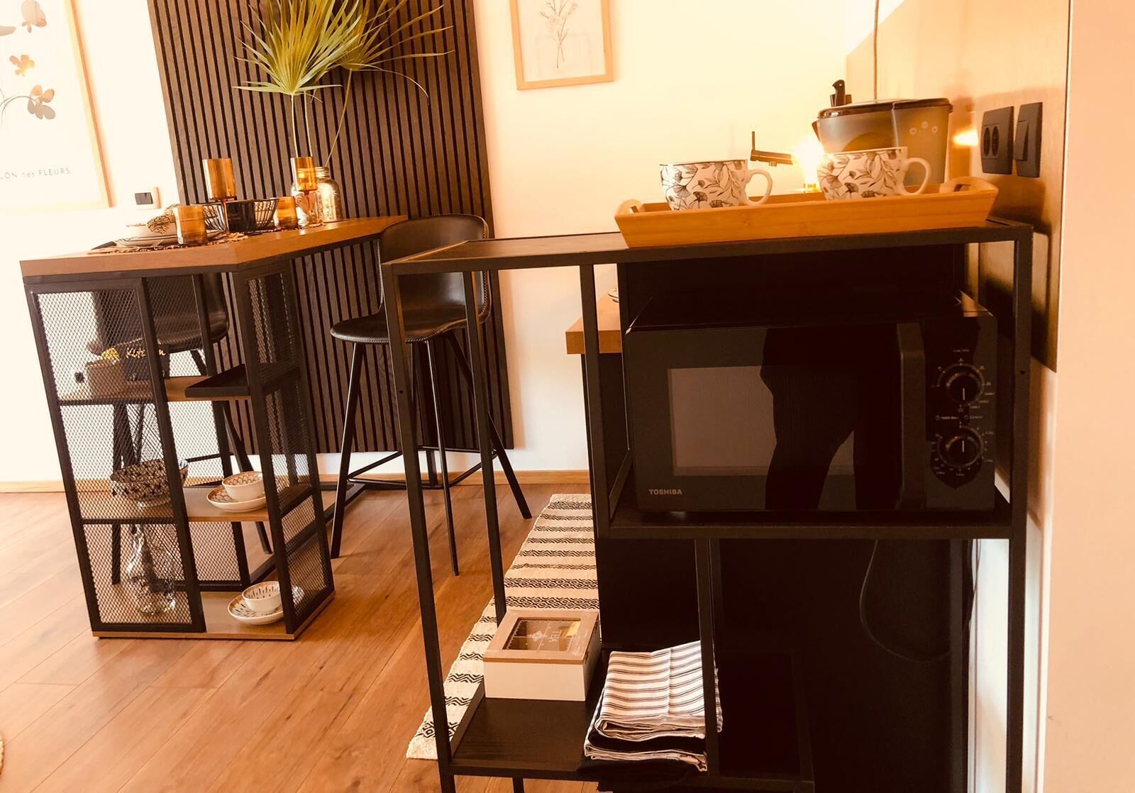 A compact kitchen with a microwave, a coffee maker, and a tray with cups and a teapot. There are two chairs and a small table with a vase and a plant.