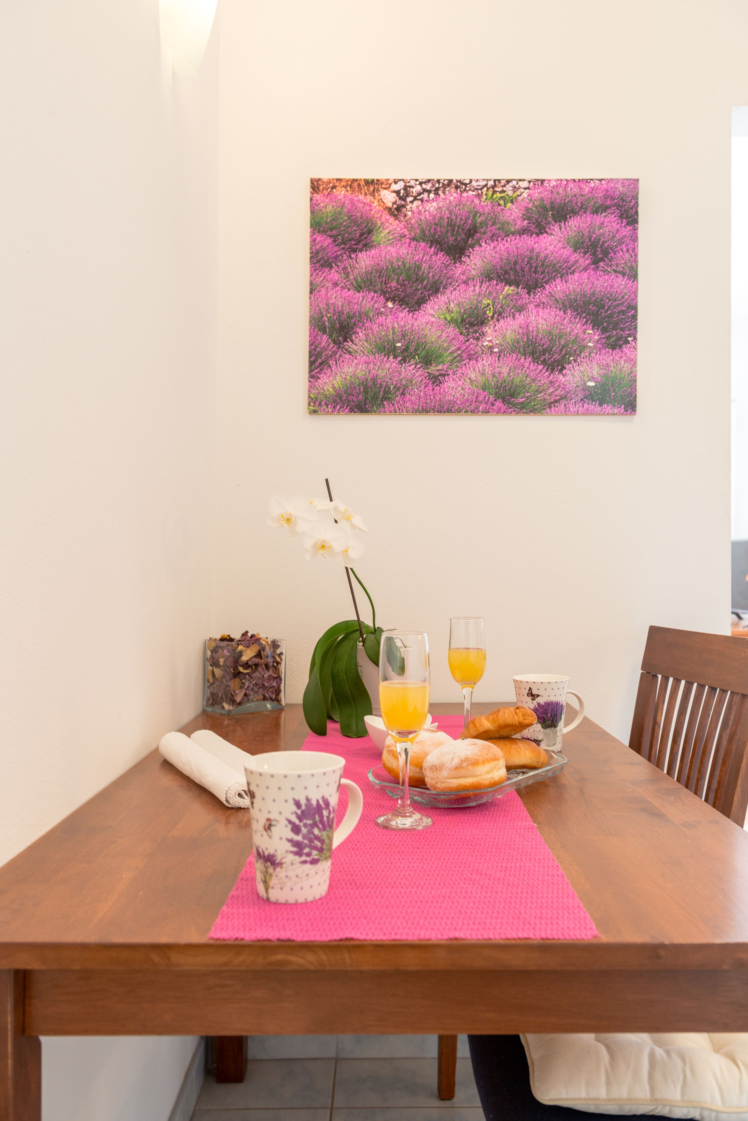 A dining table with a pink placemat, a white mug with purple flowers, a glass of orange juice, a glass of water, a plate with pastries, and a small vase with an orchid.