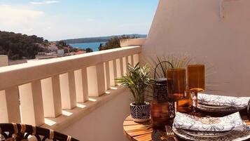 A balcony with a view of the sea, a round wooden table with a white cushion, and a potted plant.