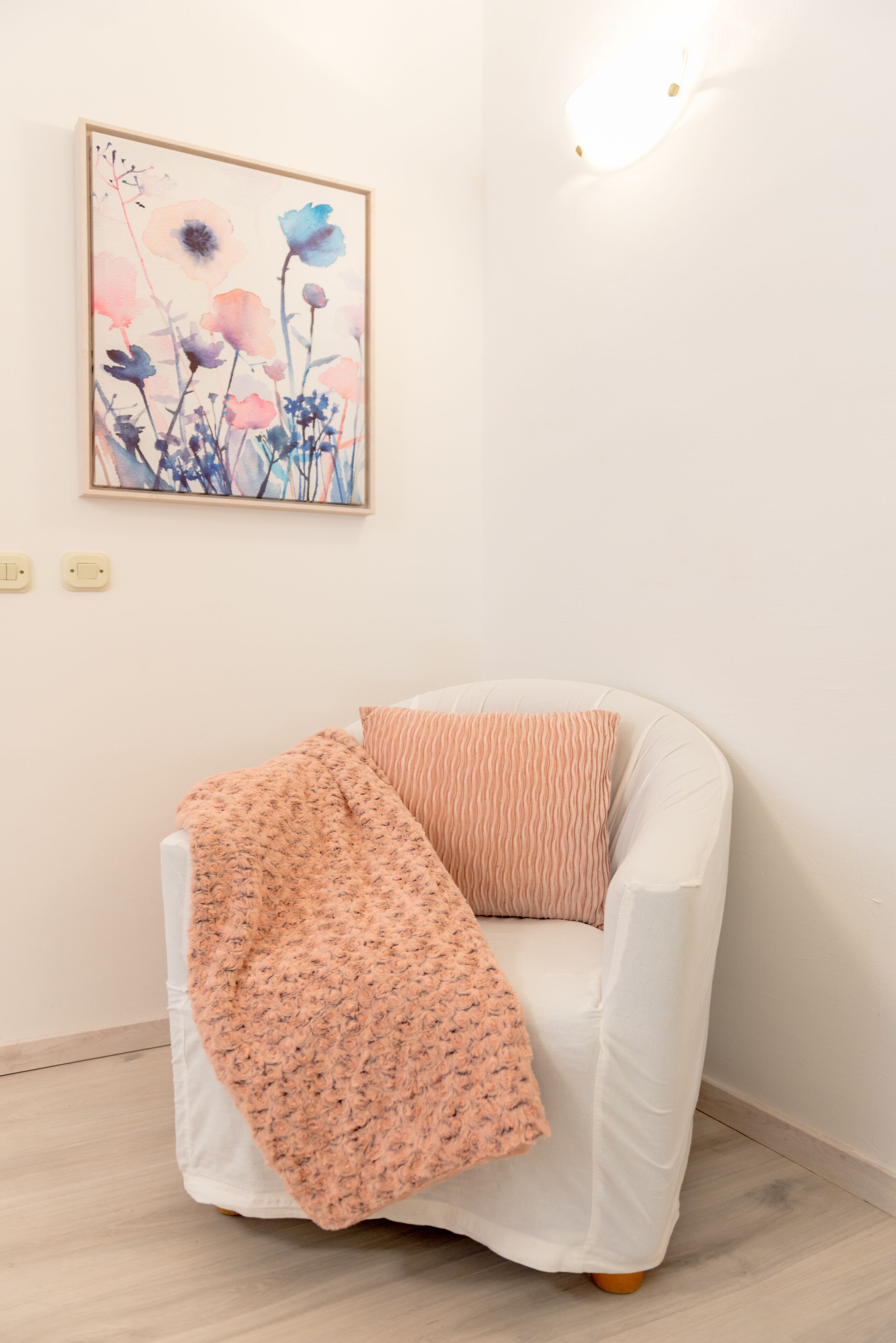 Un sillón blanco con una manta color melocotón y un cojín a rayas, ubicado frente a una pared con una obra de arte floral enmarcada y una lámpara.