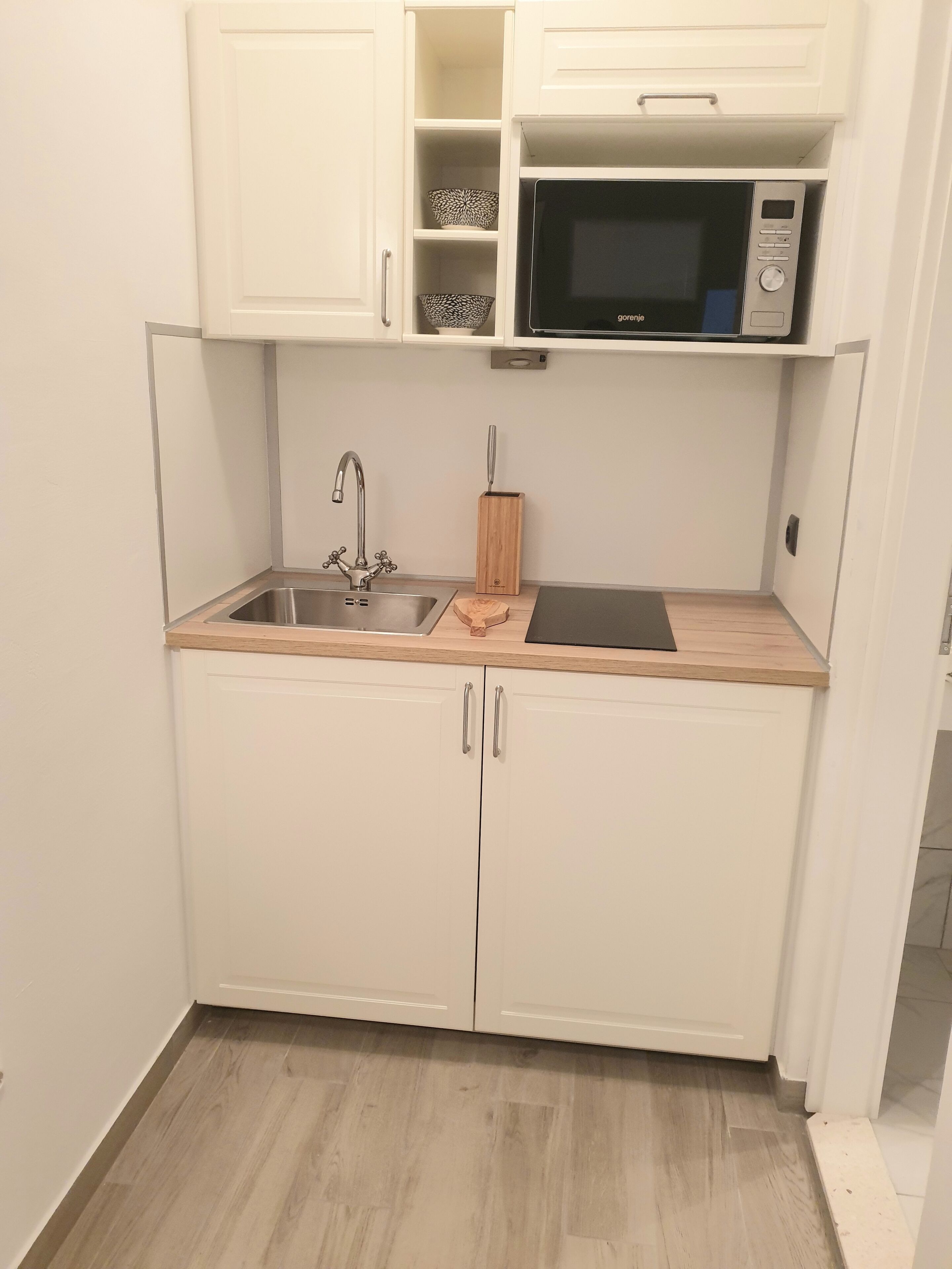 A compact kitchen with white cabinets, a stainless steel sink, a microwave, and a wooden cutting board.