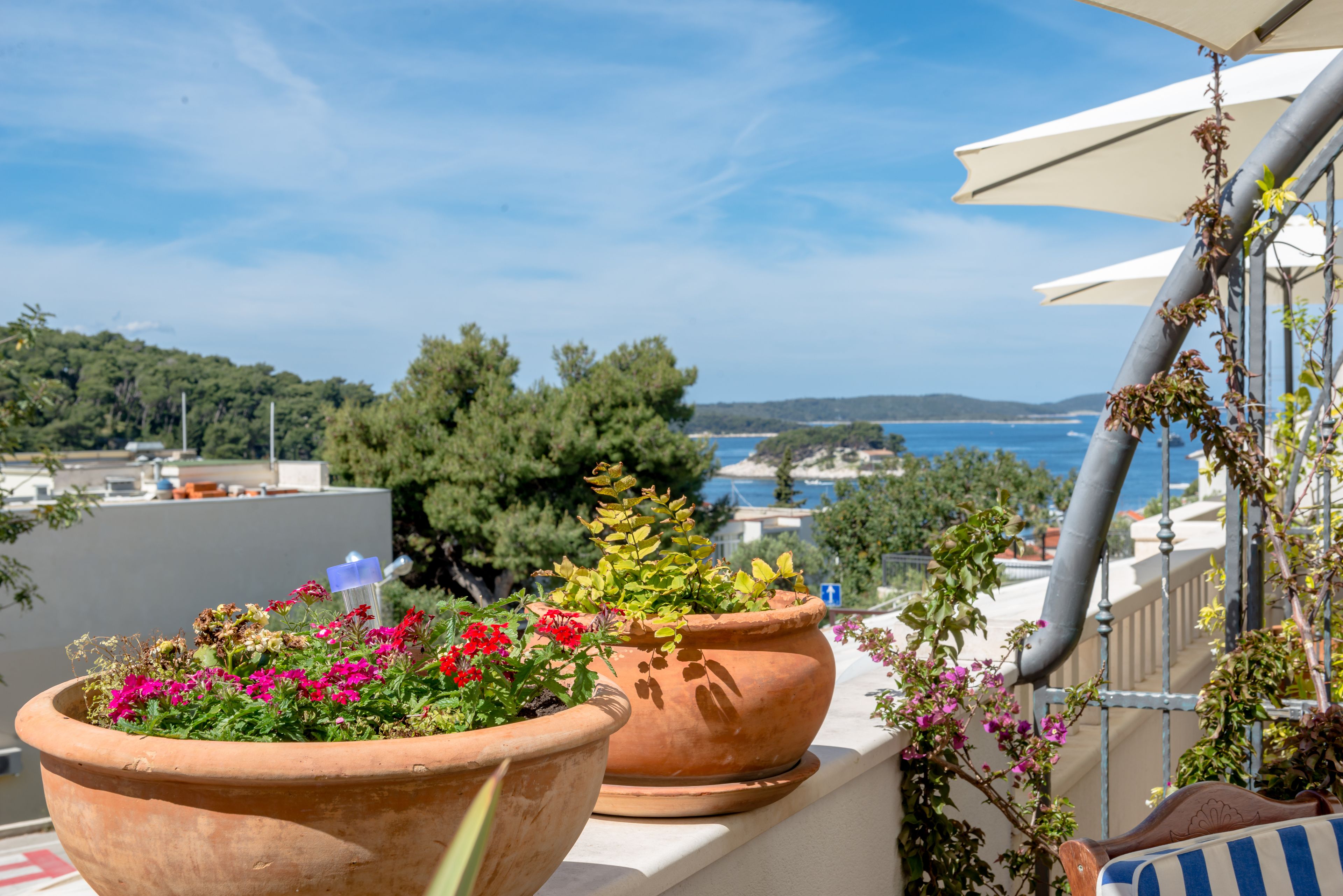 A balcony with terracotta planters containing flowers, overlooking a coastal landscape with trees and a view of the sea.
