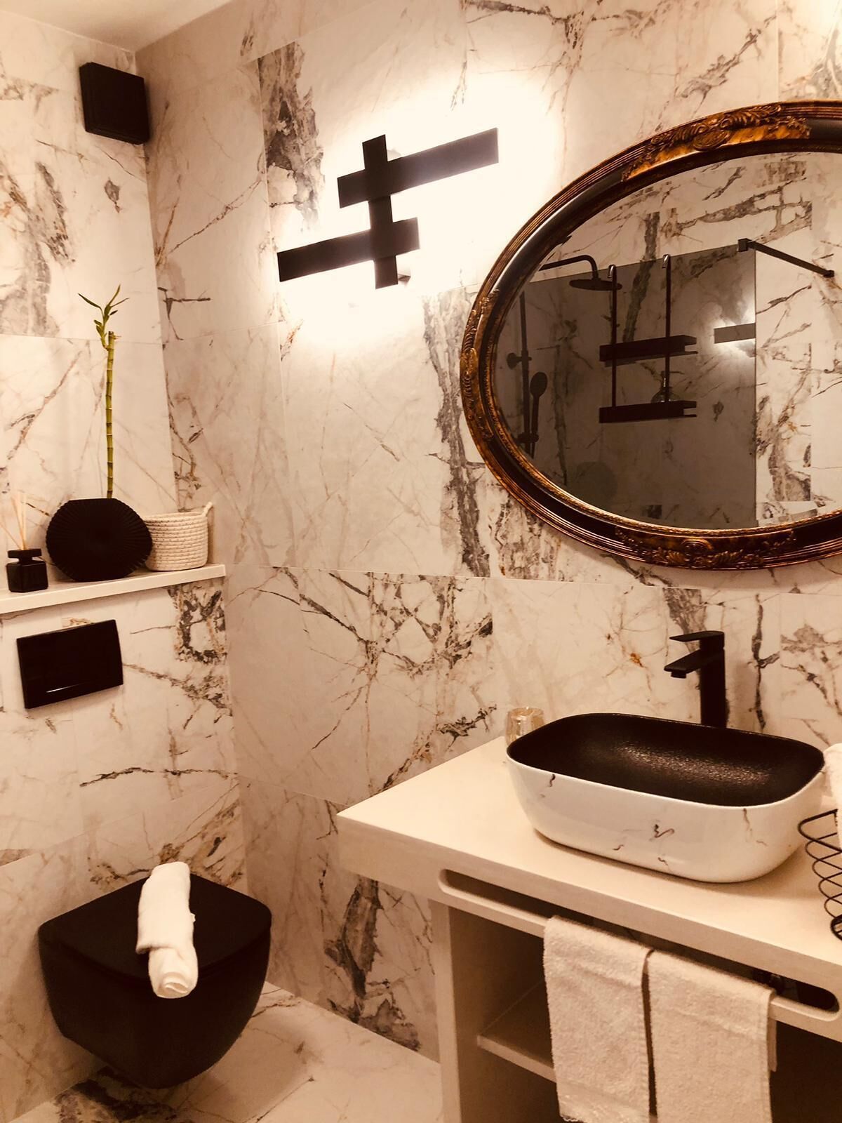 Un baño moderno con pared de mármol, lavamanos negro, un espejo grande y un inodoro negro.