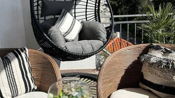 Een rieten stoel met kussens, een glazen terrarium en een geweven mand op een balkon.