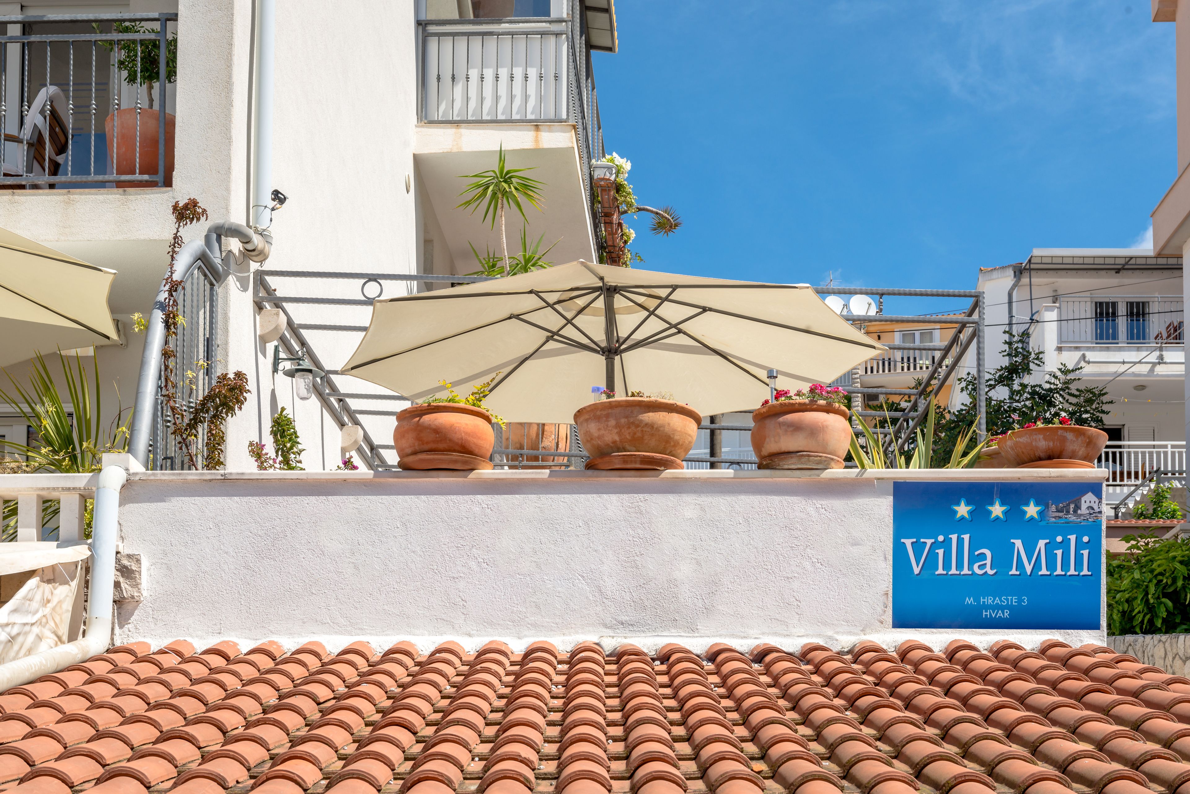 Un toit recouvert de tuiles, une enseigne indiquant « Villa Mili », et des plantes en pot sur un balcon.