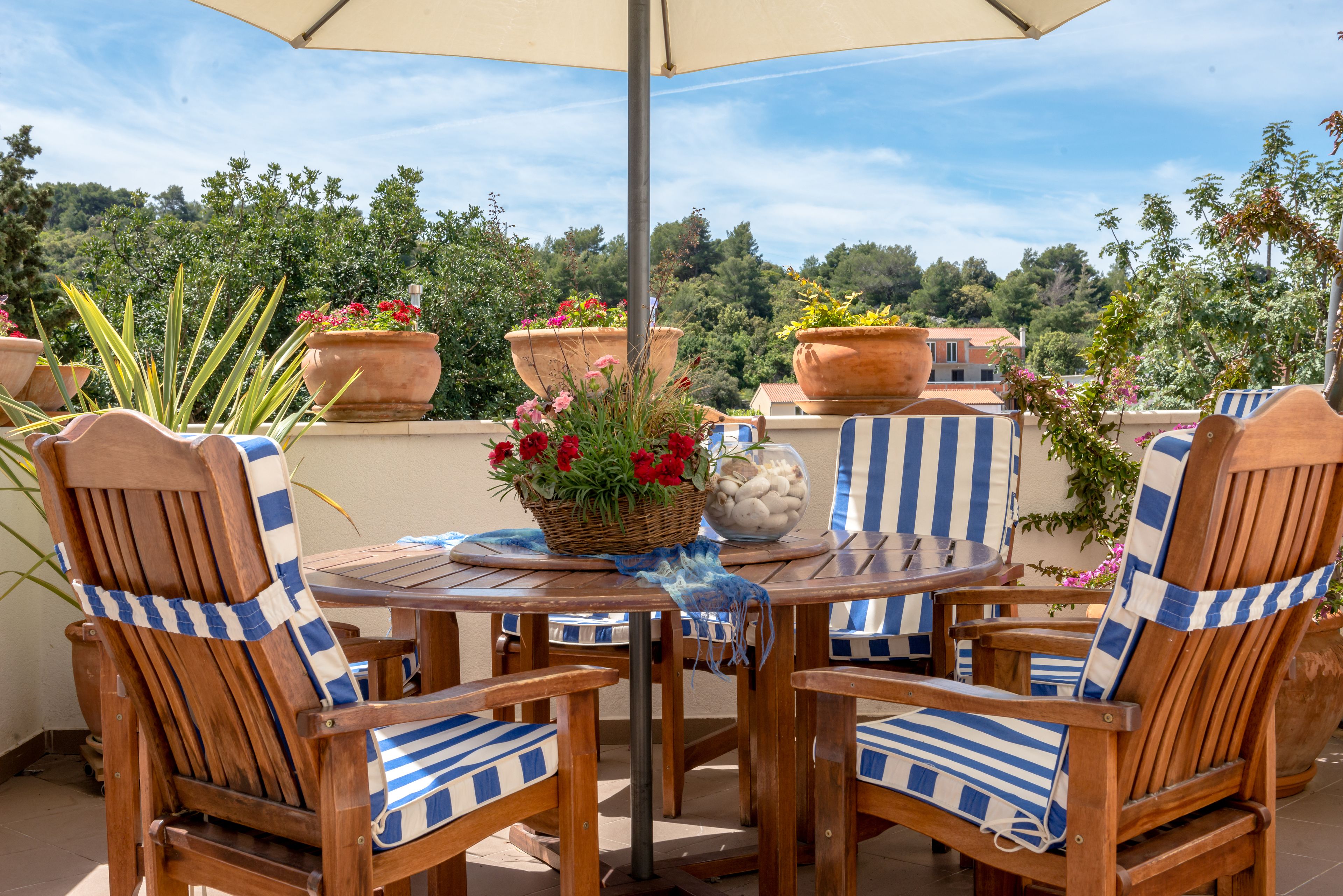 A wooden outdoor table with striped chairs, surrounded by potted plants and a basket of flowers.