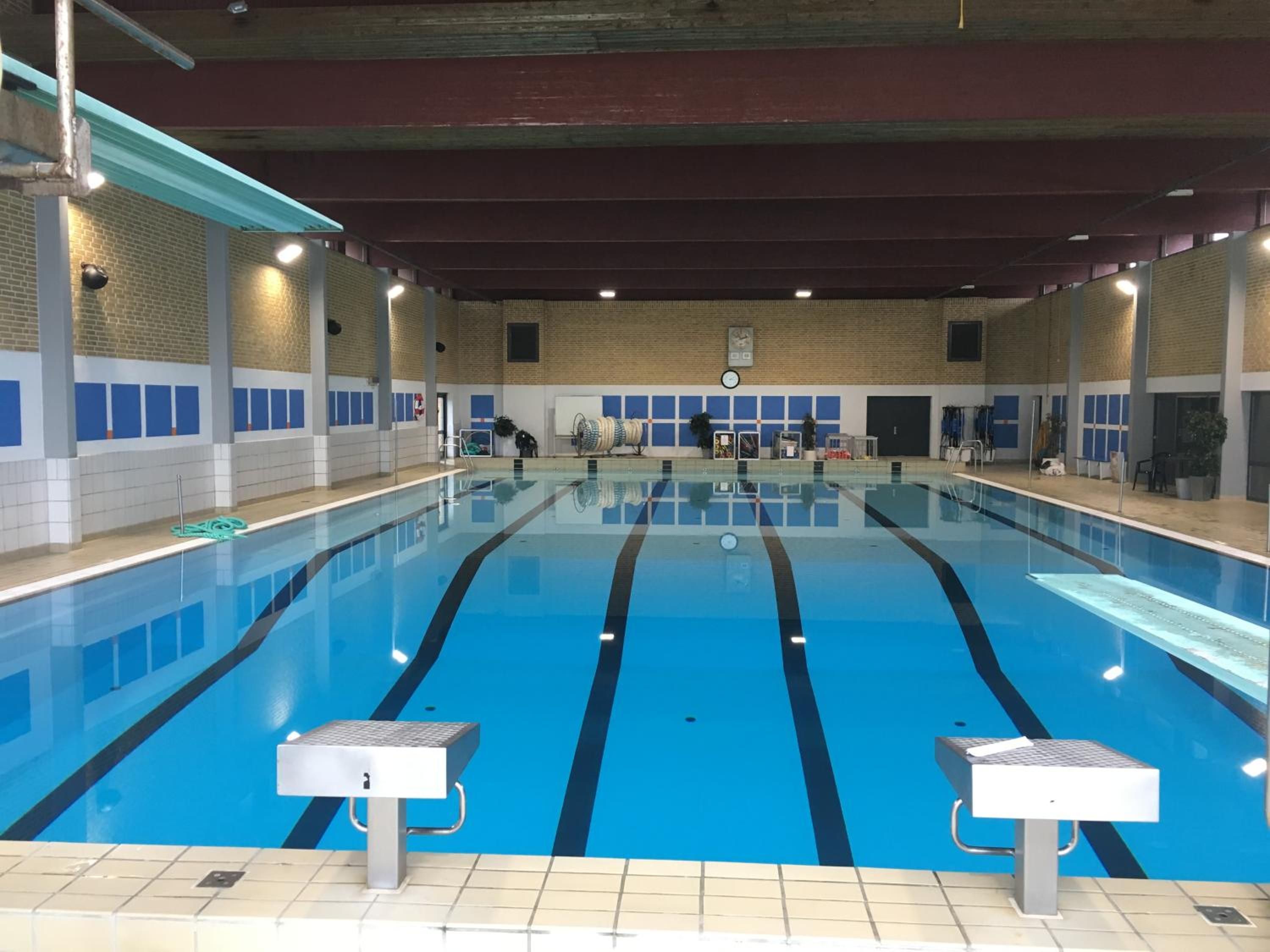 Ein Hallenbad mit deutlichen Bahnen, einer Uhr an der Wand und einem Rettungsschwimmerstuhl.