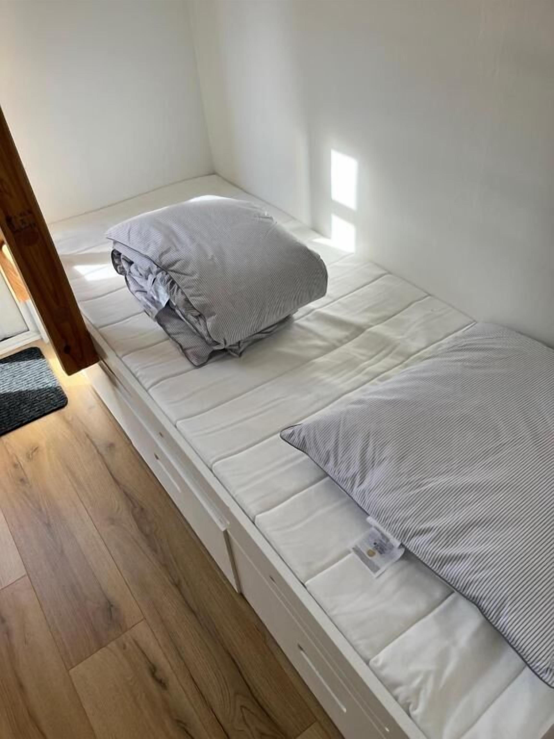 Una cama individual con colchón blanco y dos almohadas, suelo de madera y una ventana pequeña que proporciona luz natural.