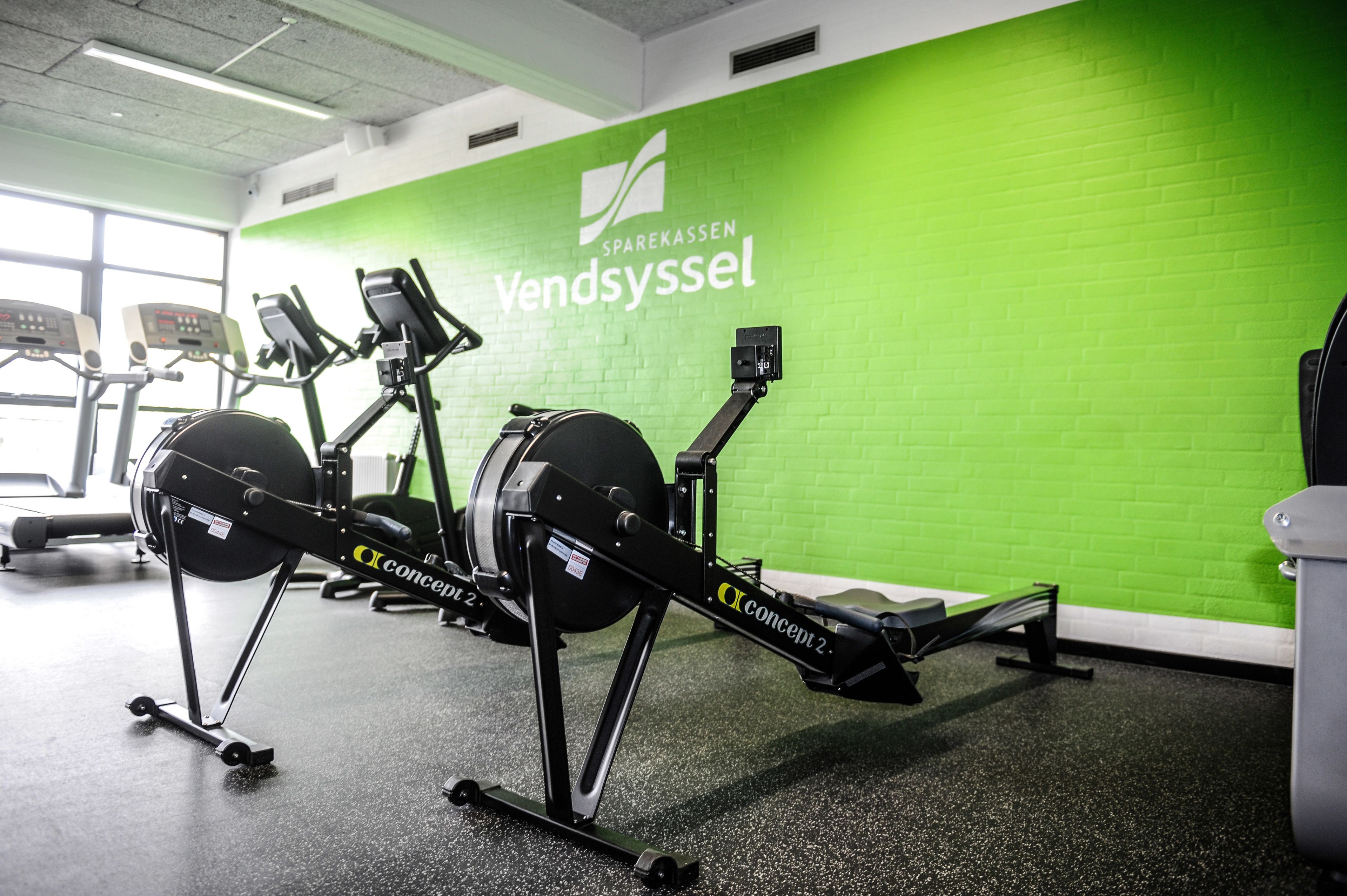 Un gimnasio con bicicletas estáticas y cintas de correr, una pared verde con el texto "SPAREKASSEN Vendsyssel" y un suelo negro.