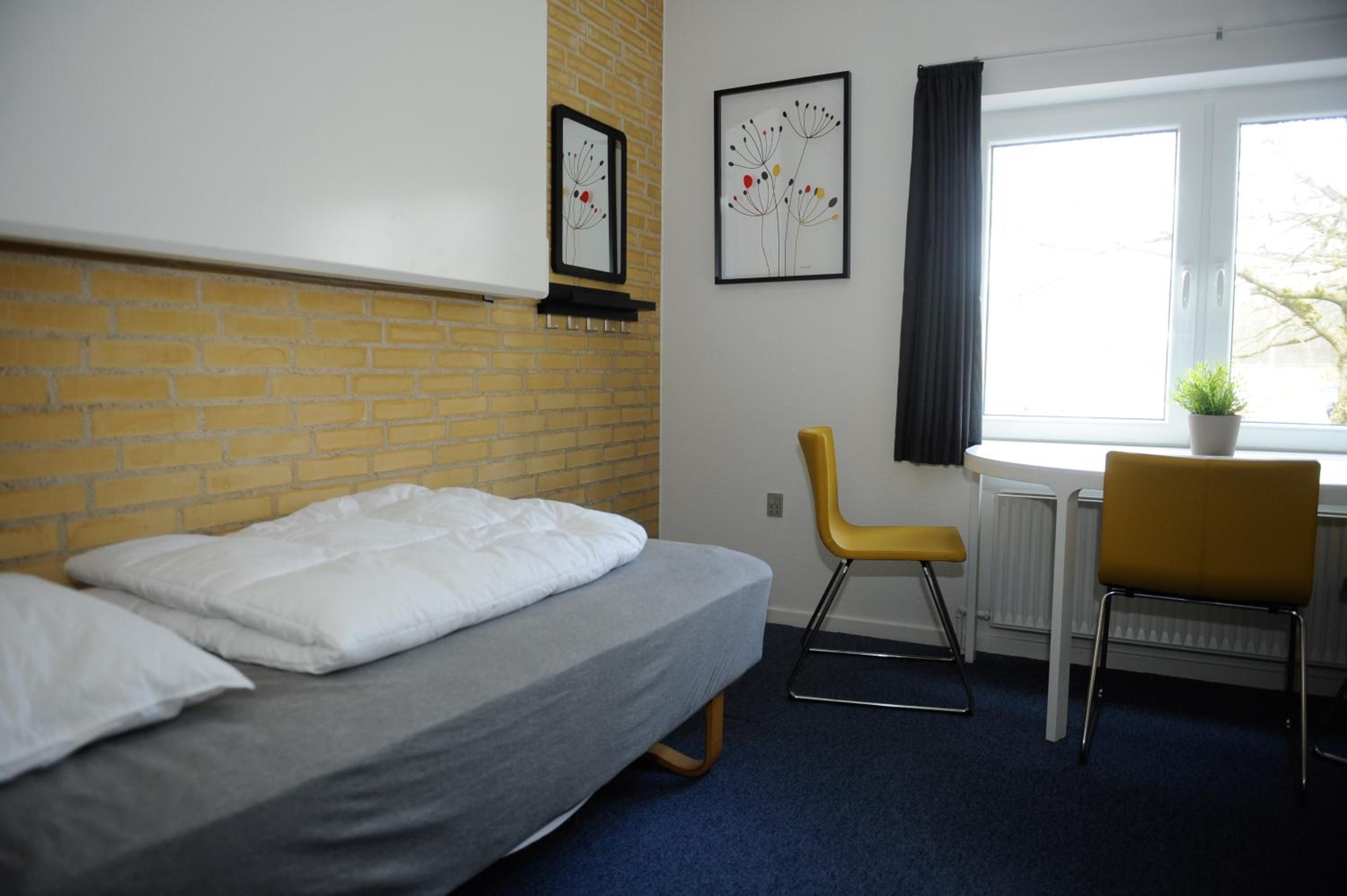 Une pièce comprenant un lit, une chaise, un bureau, une fenêtre et deux tableaux accrochés au mur.