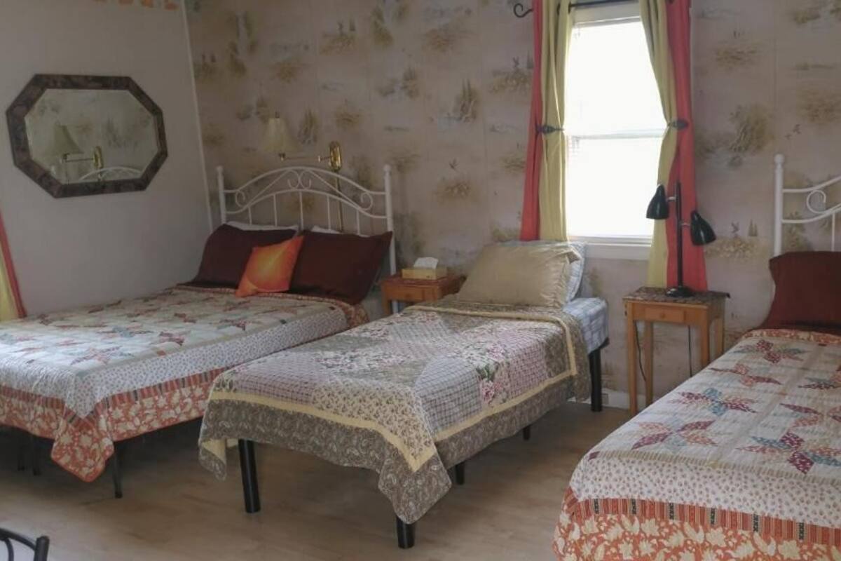A room with three single beds, each with a patterned bedspread and pillows. There is a small table with a lamp and a nightstand with a lamp. The room has floral wallpaper and a window with curtains.