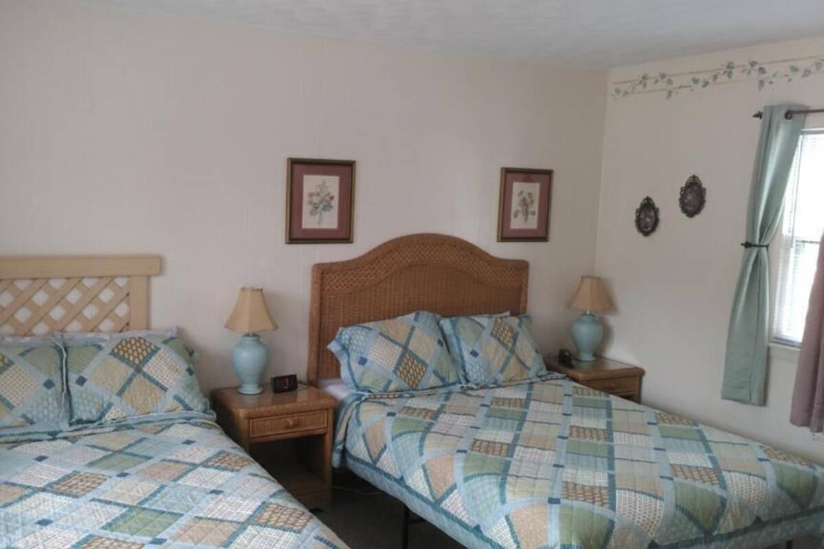 A bedroom with two beds, each with a headboard and nightstands. There are framed pictures on the wall and a window with curtains.