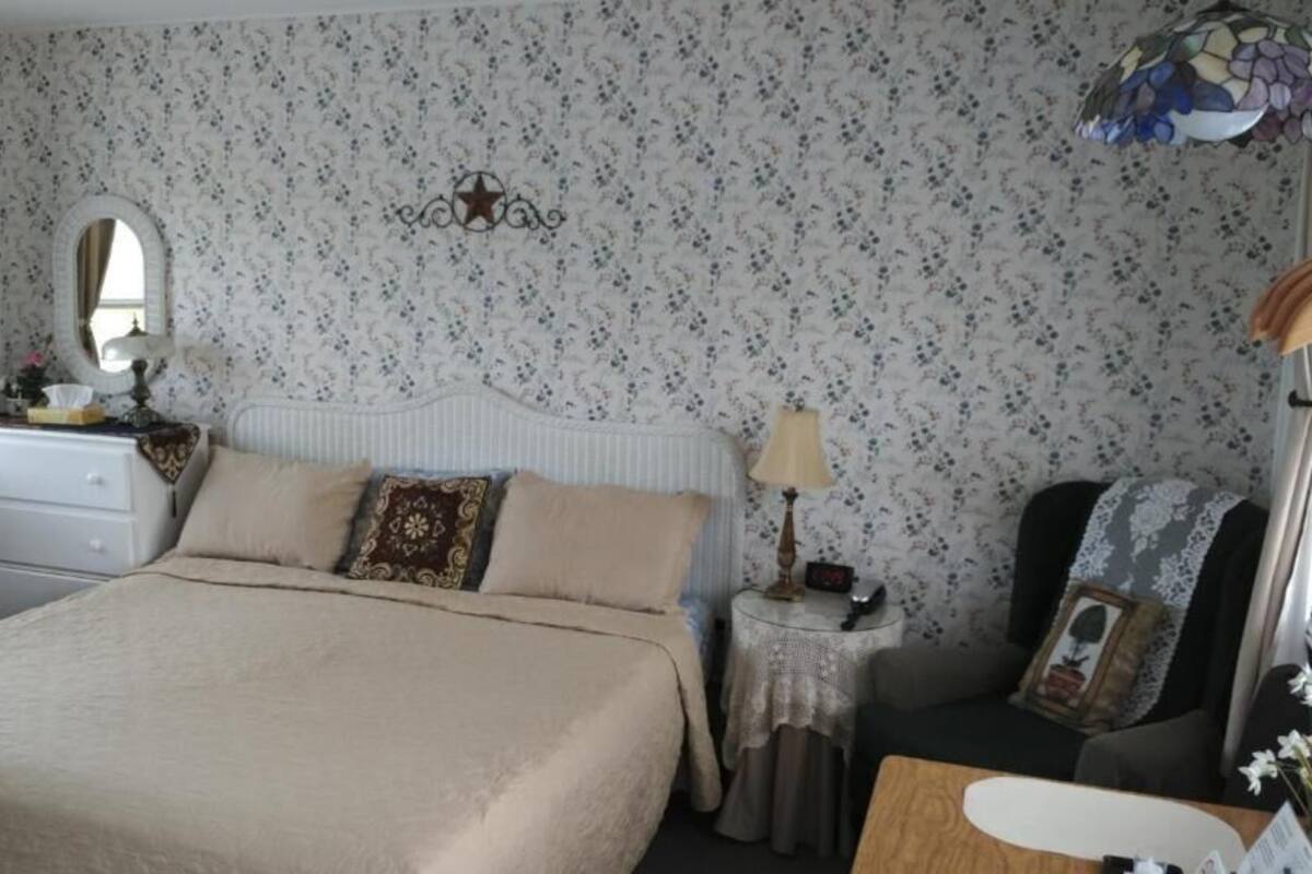 A bedroom with a large bed, a dresser, a nightstand, a chair, and a window with curtains.