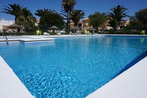 En pool med kristallklart vatten, omgiven av solstolar och parasoller, palmer och byggnader.