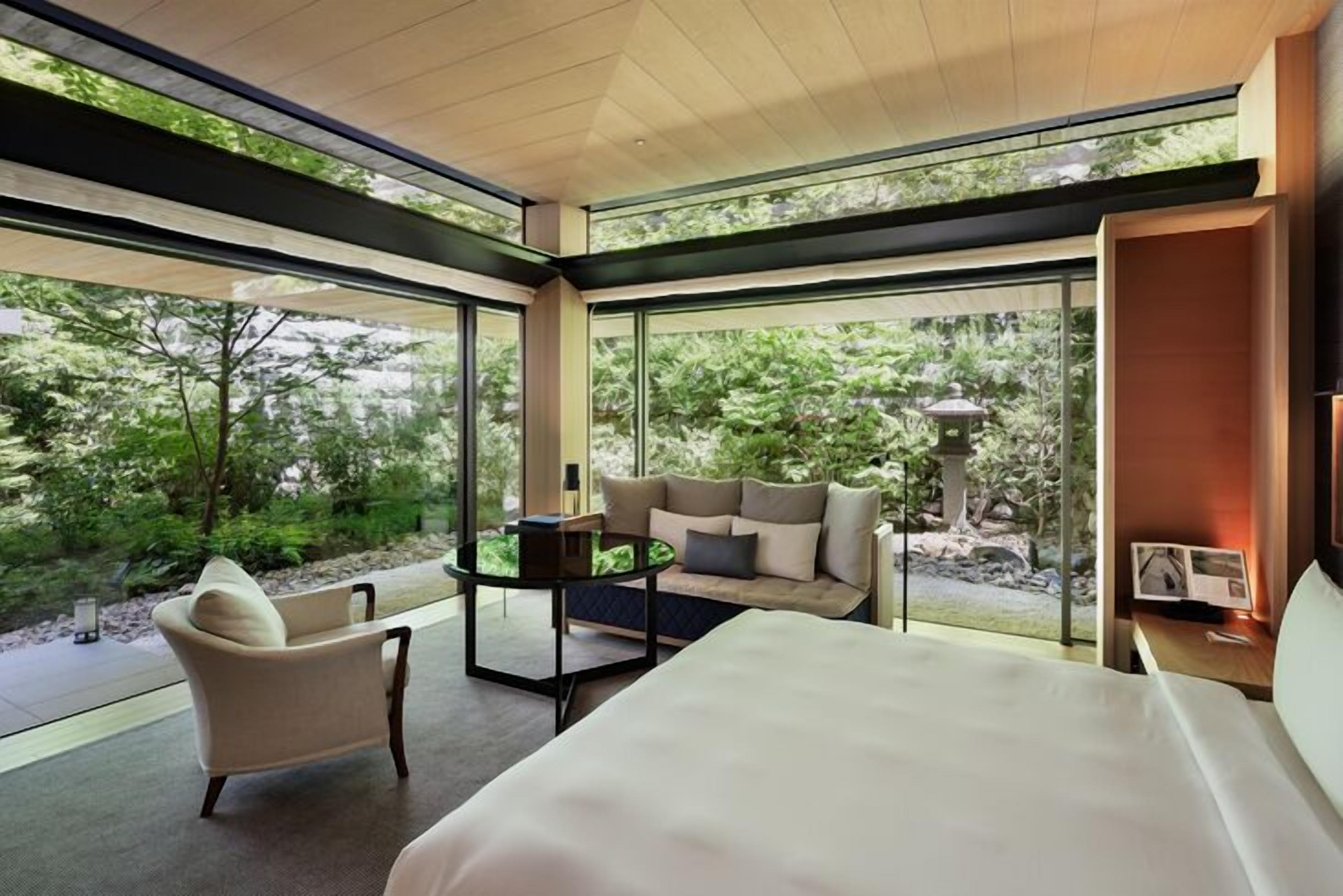 A modern hotel room with a large bed, a sofa, a chair, and a small table. There is a view of greenery outside through large windows.