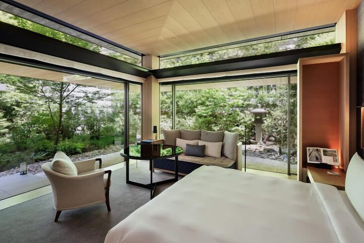 Habitación de hotel moderna con una cama grande, un sofá, una silla y una mesita. Se ve follaje verde a través de amplias ventanas.