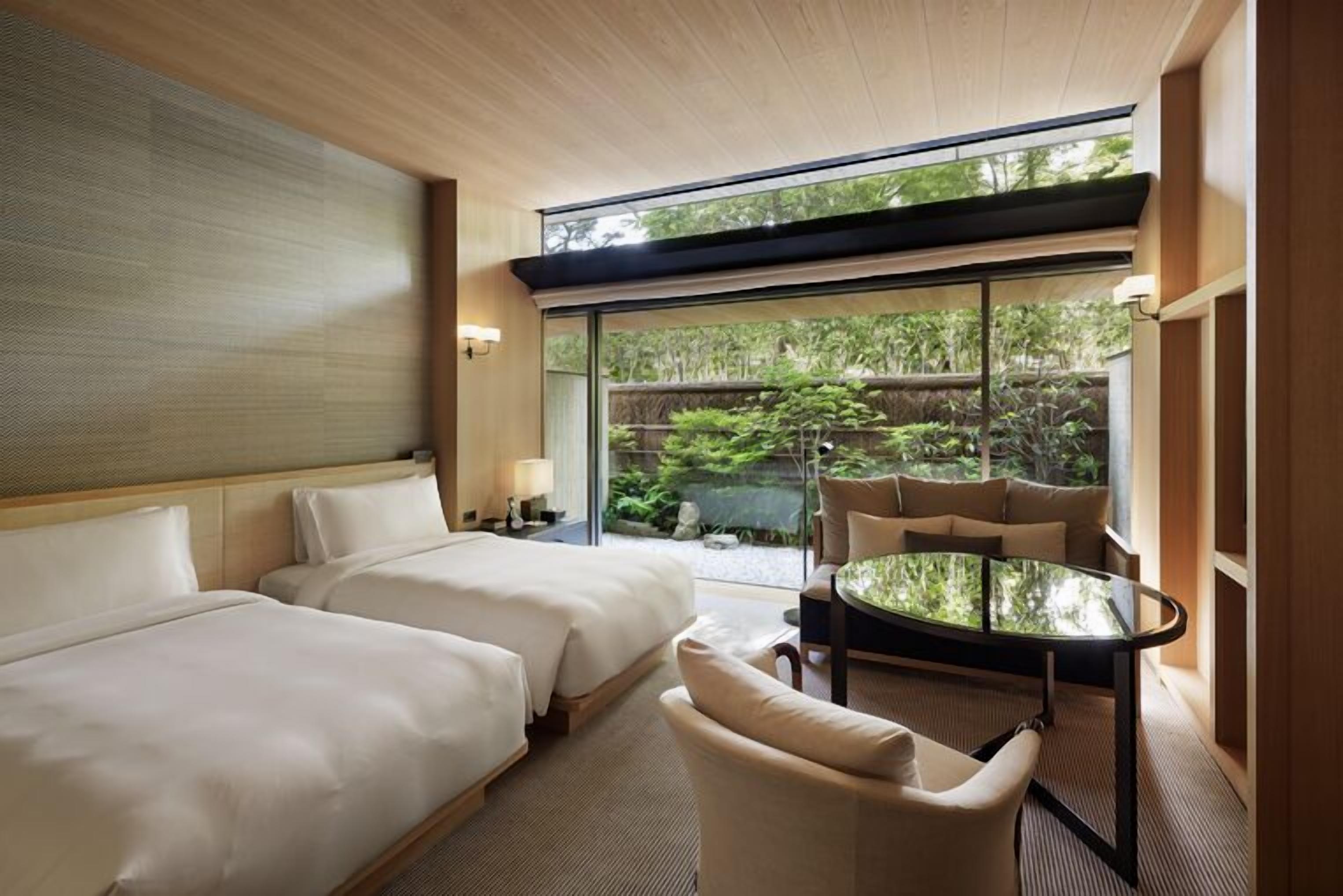 A hotel room with two beds, a glass table, and a view of greenery outside.