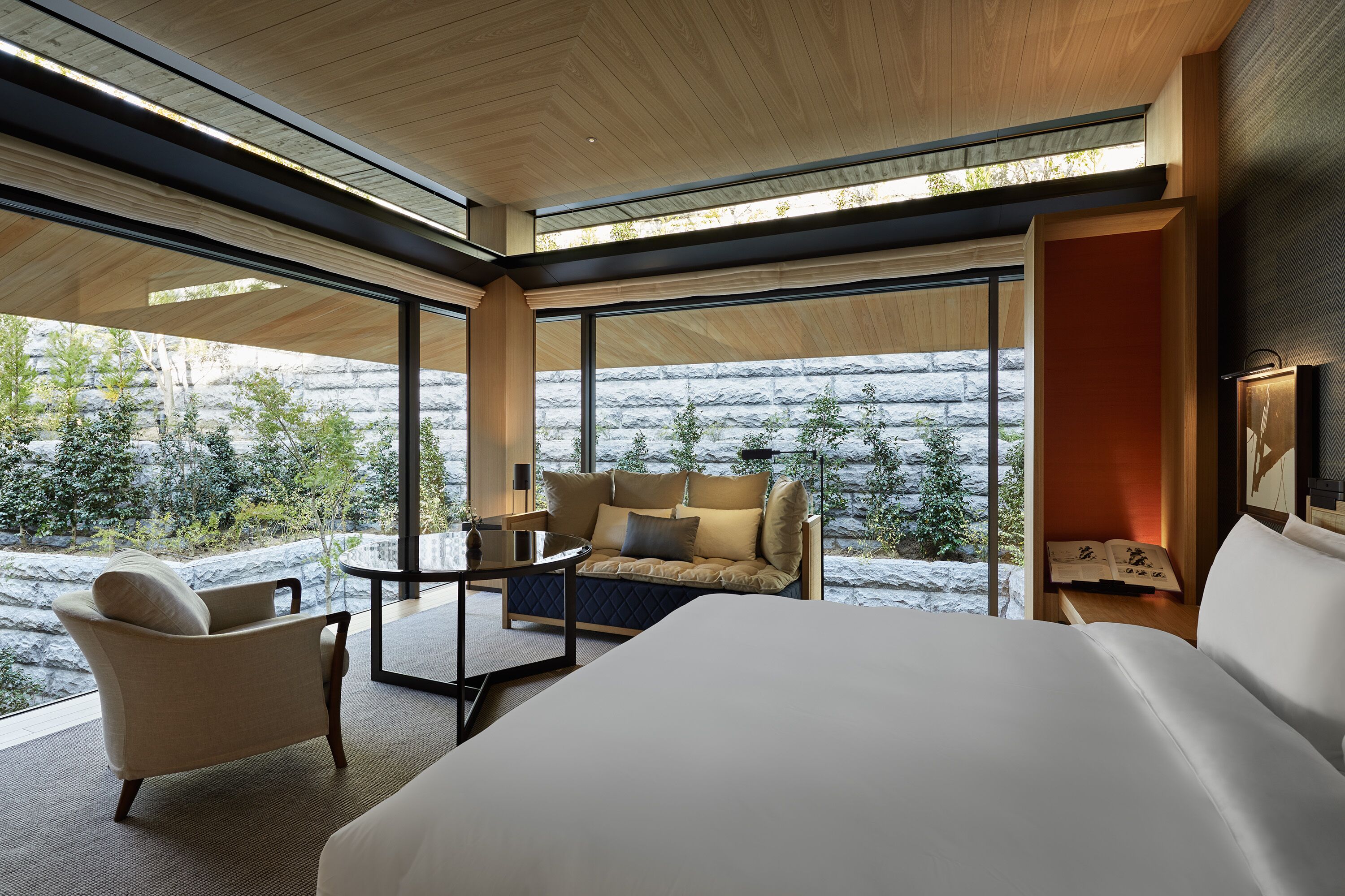 A modern hotel room with a large bed, a sofa, a small table, and a chair. The room has large windows offering a view of a rocky landscape.