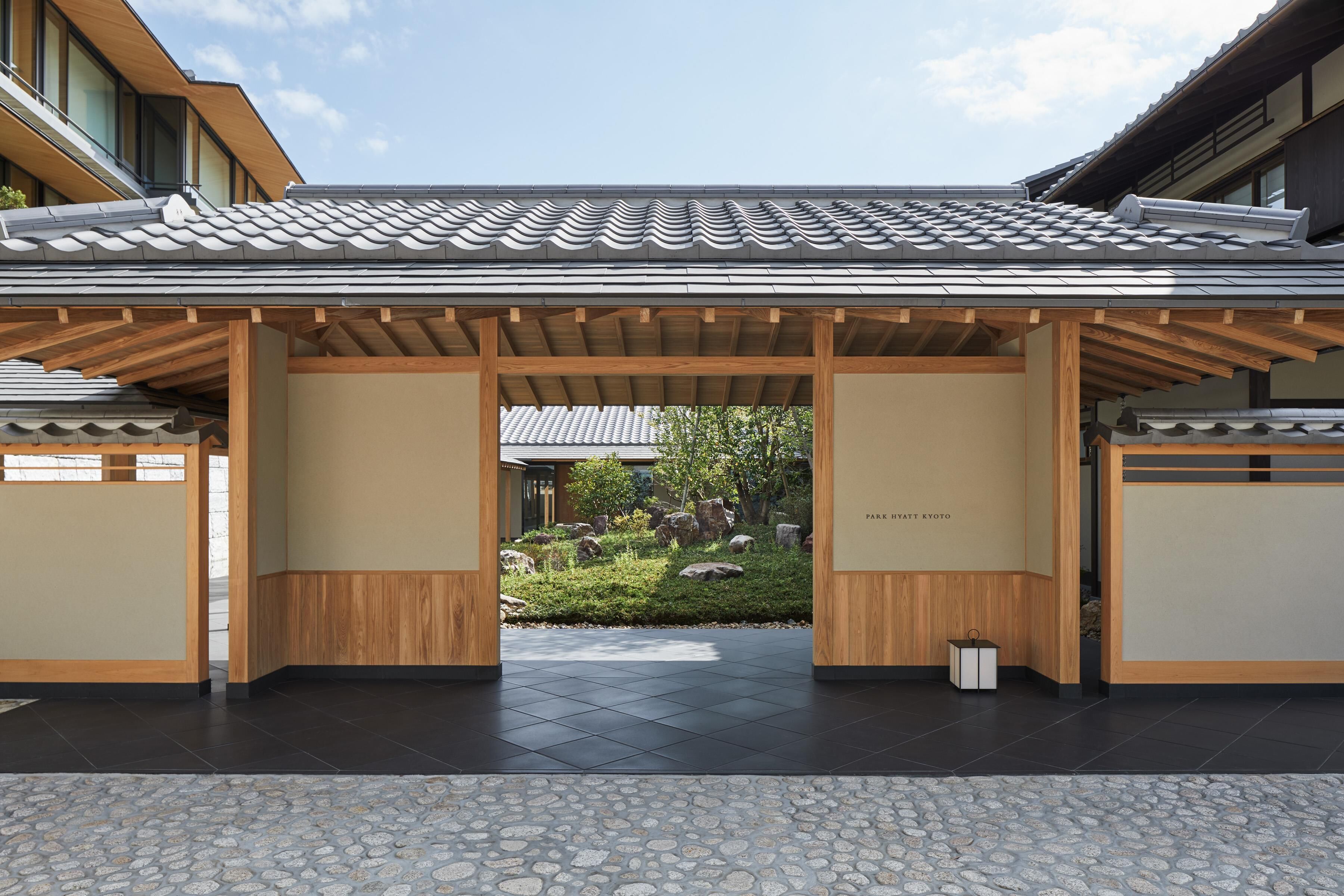 A traditional Japanese-style building with a tiled roof and wooden beams, a stone-tiled entrance, and a small garden with rocks and greenery.