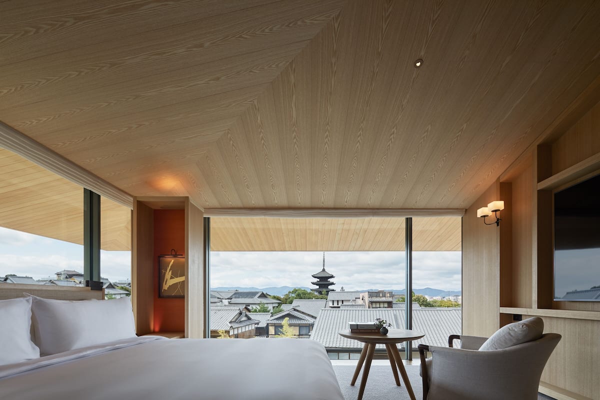 Ein modernes Hotelzimmer mit einem großen Bett, einem kleinen Tisch und einem Stuhl. Das Zimmer verfügt über große Fenster mit Blick auf traditionelle Gebäude und eine Pagode.