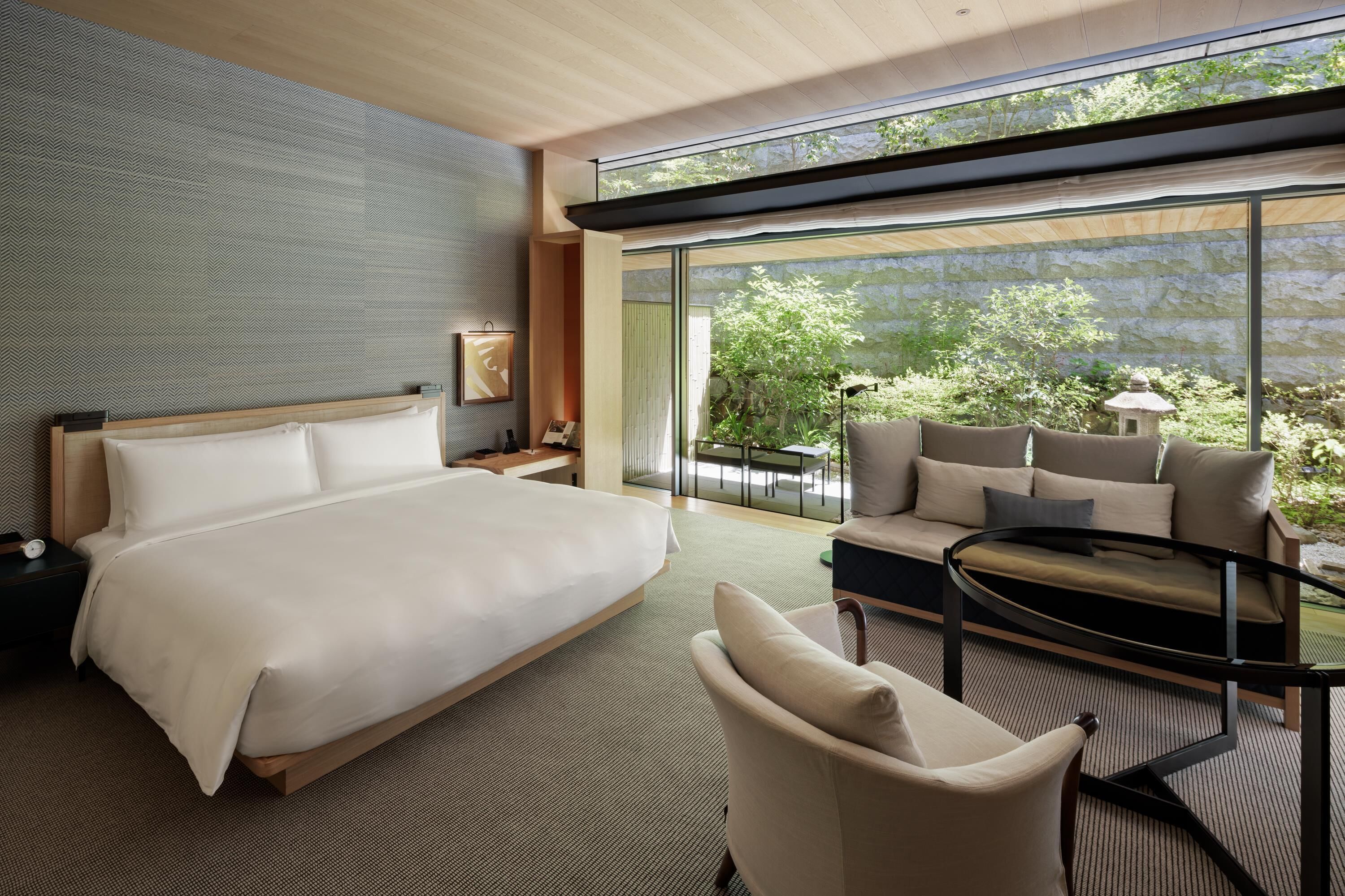 A modern hotel room with a large bed, a sofa, a small table, and a view of greenery through large windows.